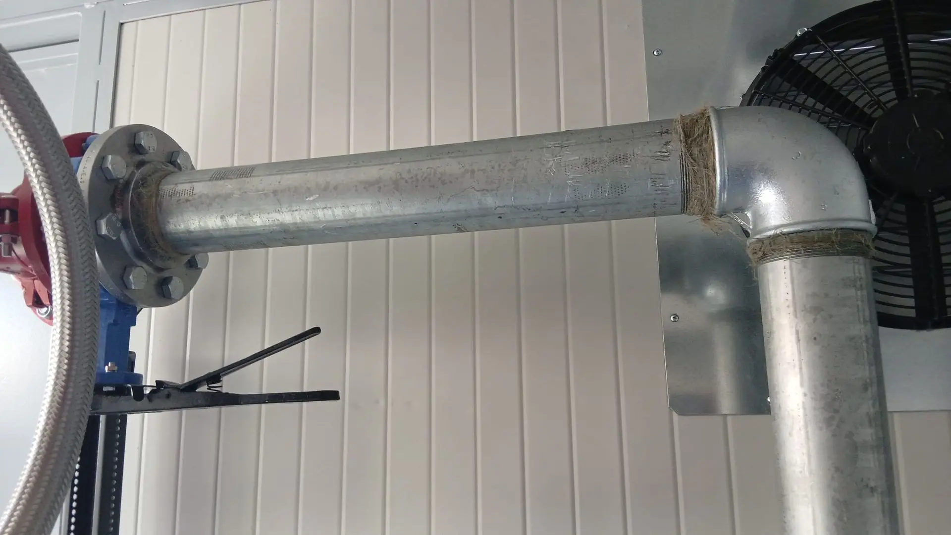 Tubazione industriale in acciaio zincato con flangia imbullonata, valvole e ventilatore in un locale tecnico.